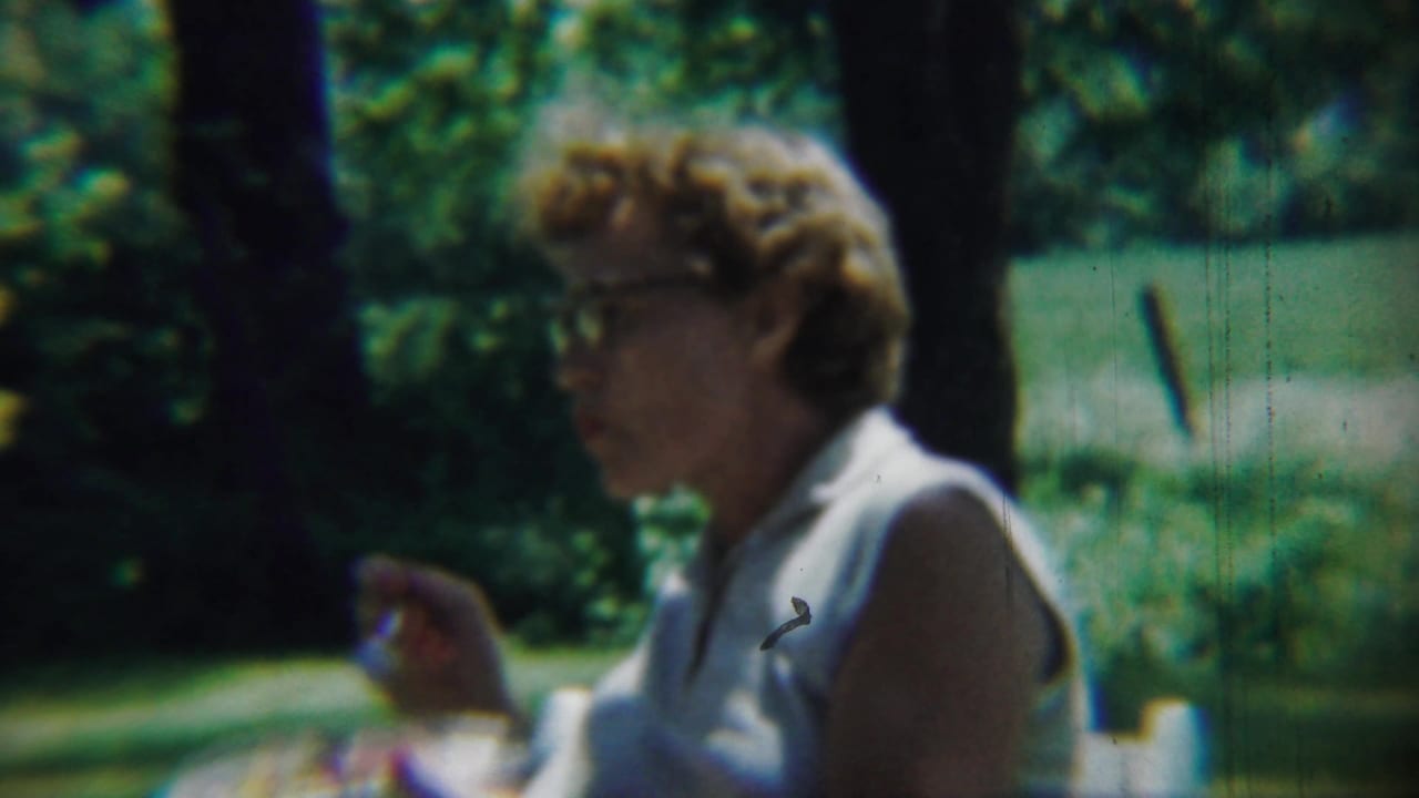 Woman in glasses sits outdoors holding food or a utensil near a sunlit lawn.