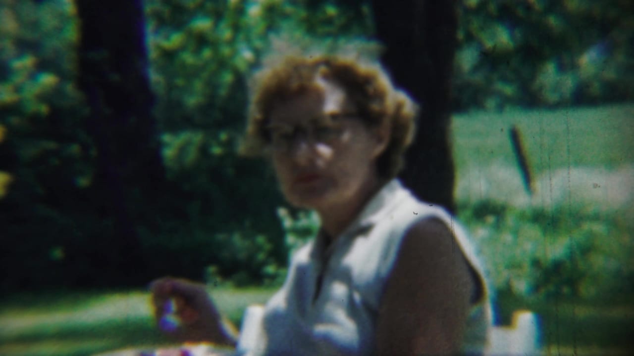 Woman with glasses turns toward the camera while seated outdoors in sunlight.