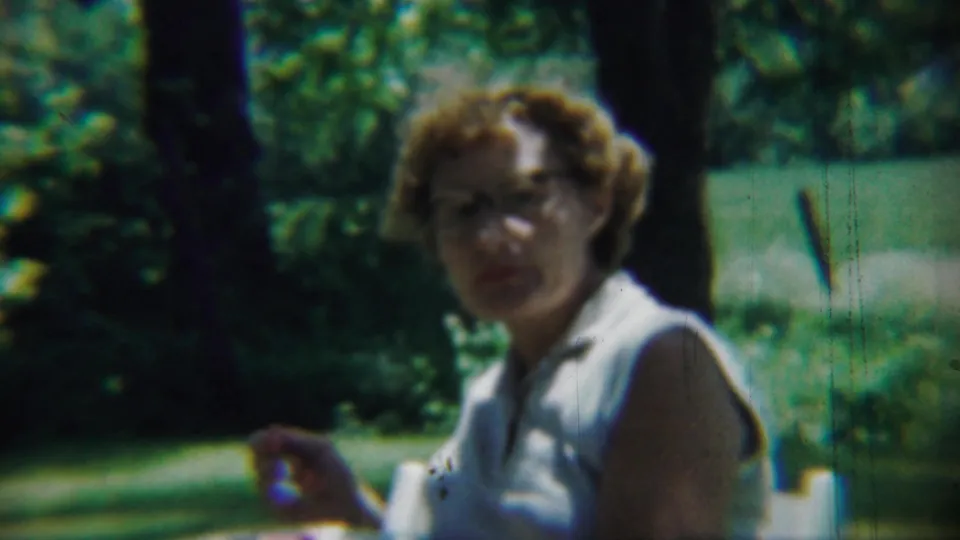 The woman faces the camera at the end of the short 1959 Chadron, Nebraska park meal clip, with lawn and trees behind her.