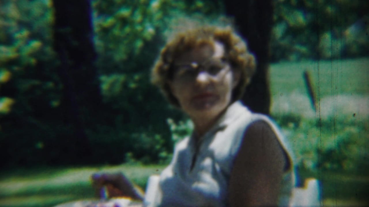 In 1959 Chadron, Nebraska footage, an adult woman with glasses appears outdoors in a white sleeveless shirt beside green foliage.