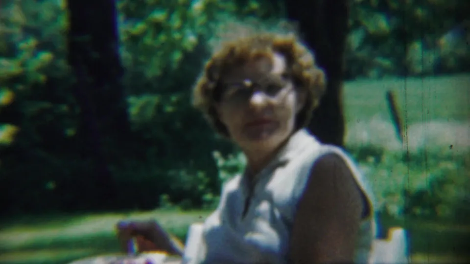 In 1959 Chadron, Nebraska footage, an adult woman with glasses appears outdoors in a white sleeveless shirt beside green foliage.