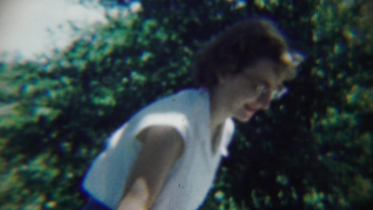 Adult woman in a white sleeveless shirt leans forward with dense greenery behind her.