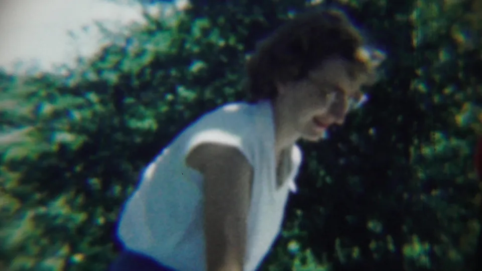 In the 1959 home-movie frame, the woman bends slightly downward near dense garden foliage, partly blurred by handheld 8mm motion.