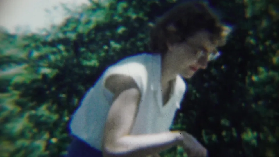 The woman looks down at the edge of a garden in the Chadron, Nebraska footage, with green shrubs and lawn behind her.