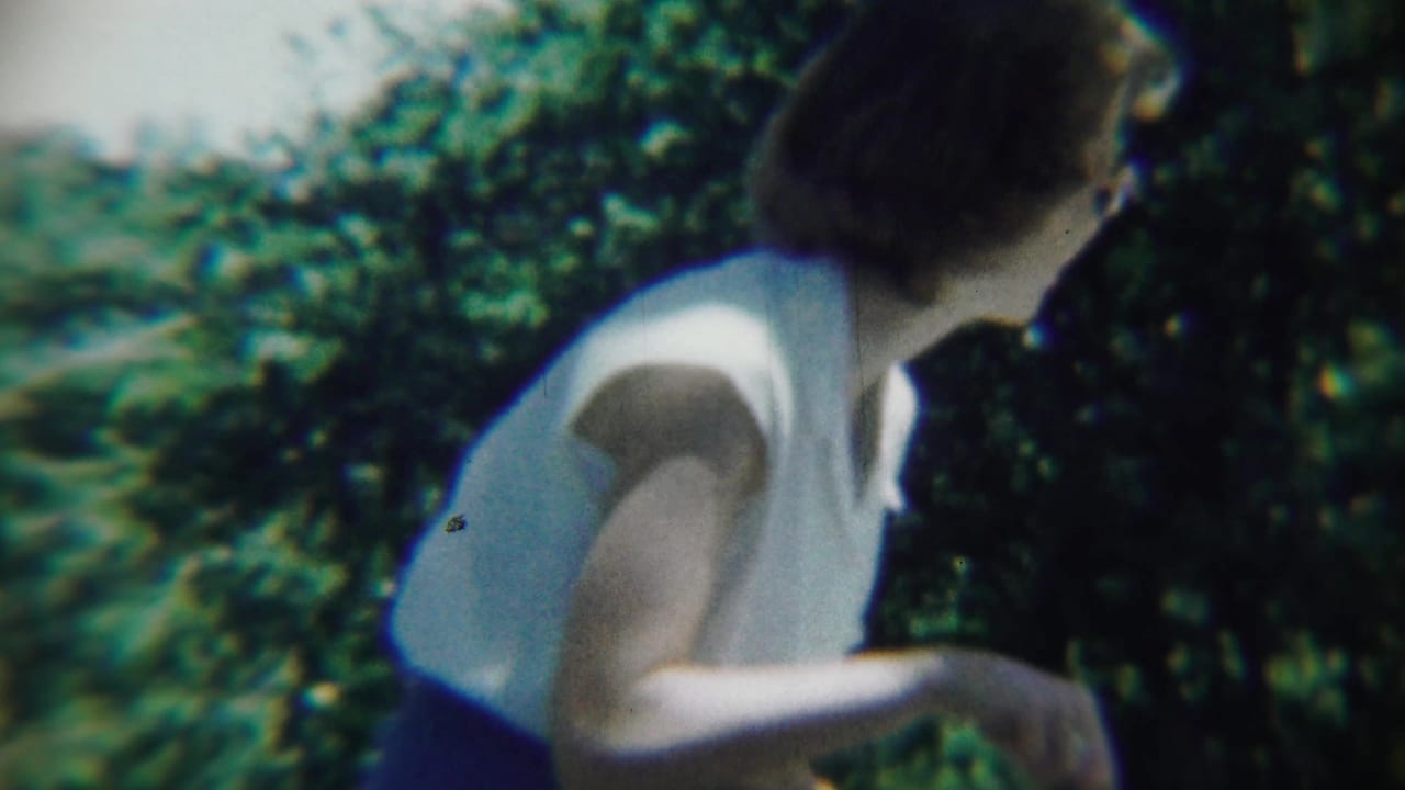 Woman in a white sleeveless top bends beside dense garden foliage.