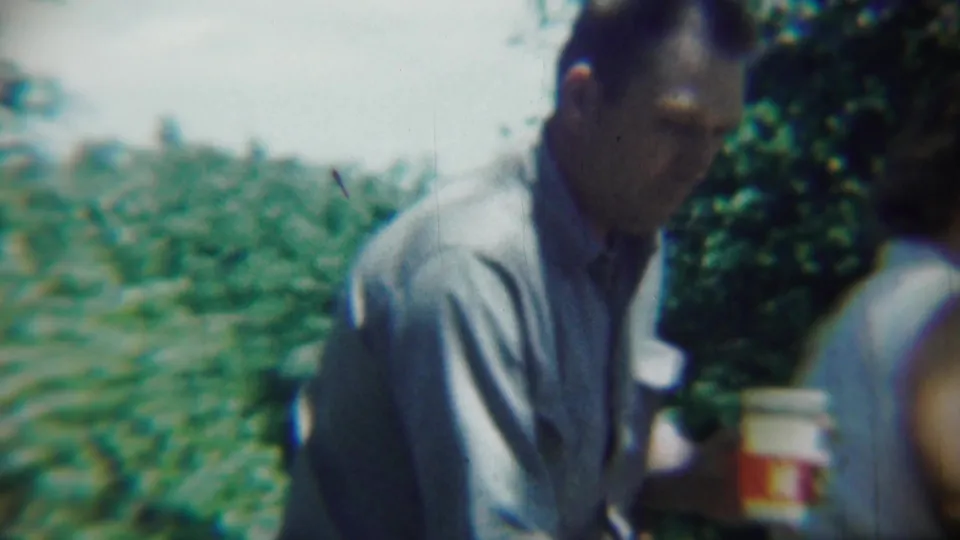 A man in a light shirt holds a cup near the woman at the edge of a green garden in Chadron, Nebraska home-movie footage.