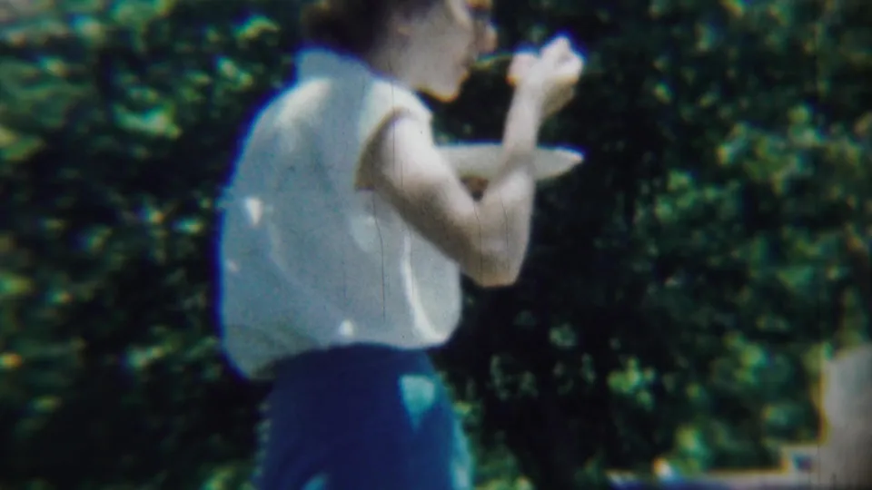 1959 Chadron, Nebraska frame showing a woman in profile eating from a paper plate amid leafy outdoor surroundings.