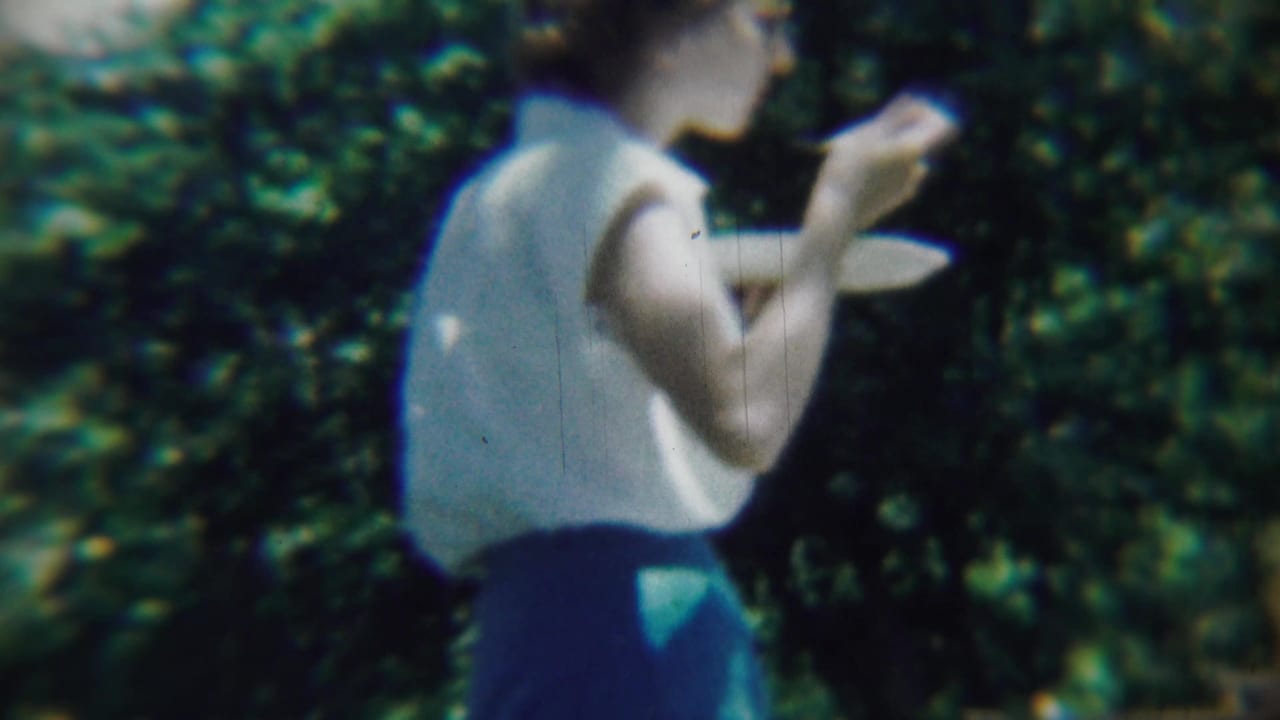 Woman in a sleeveless light top holds cake outdoors beside leafy trees.