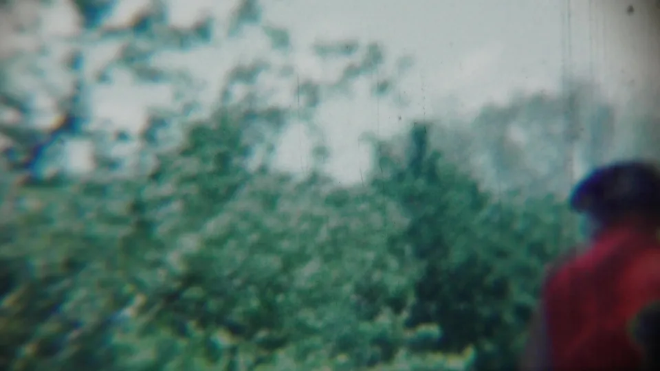 The 1959 Chadron home-movie frame shows soft garden foliage with a partially visible person in red at the right edge.