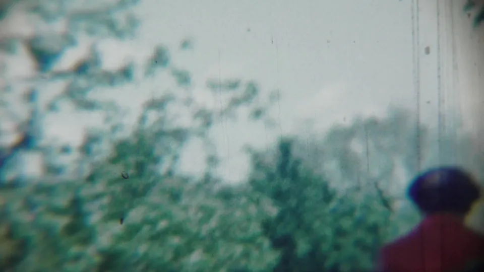 In 1959 Chadron, Nebraska home-movie footage, an adult woman in red walks away through a soft, leafy wooded area.
