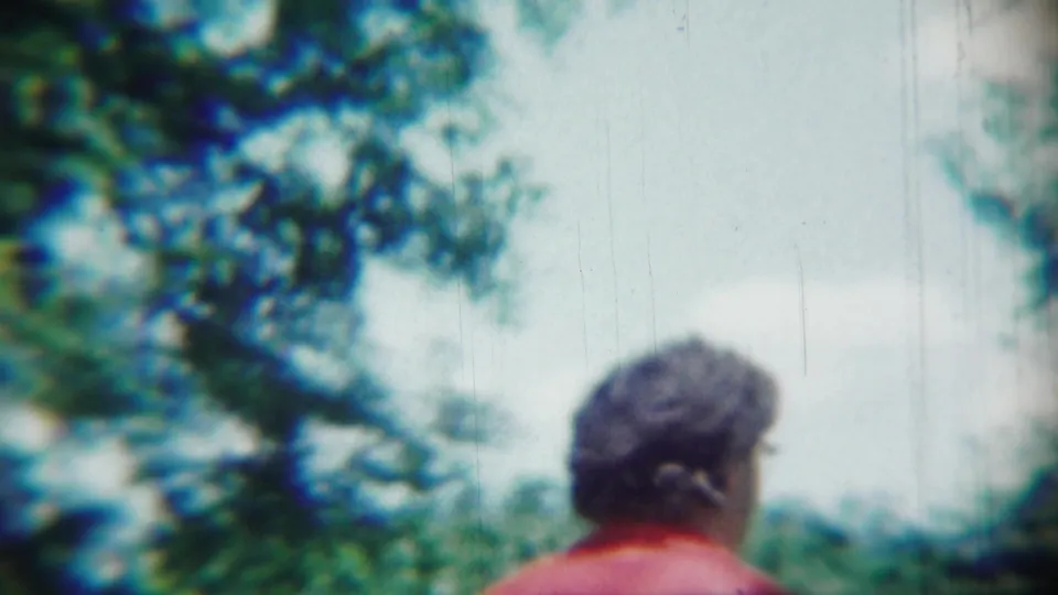 The woman in red is shown from behind near trees and bright sky, continuing the candid 1959 Chadron home-movie walk.
