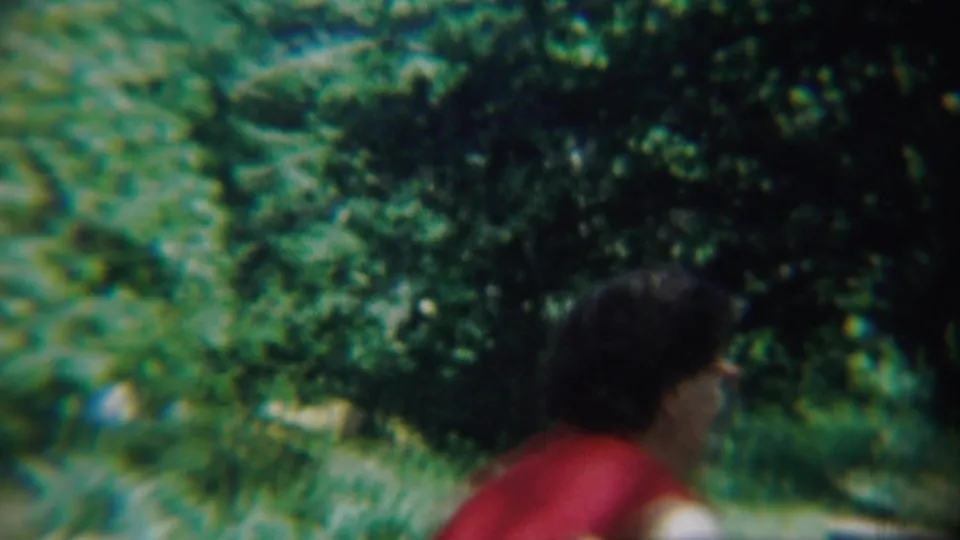 1959 Chadron, Nebraska frame showing a dark-haired person in a red shirt near bushes and trees in soft-focus 8mm color.
