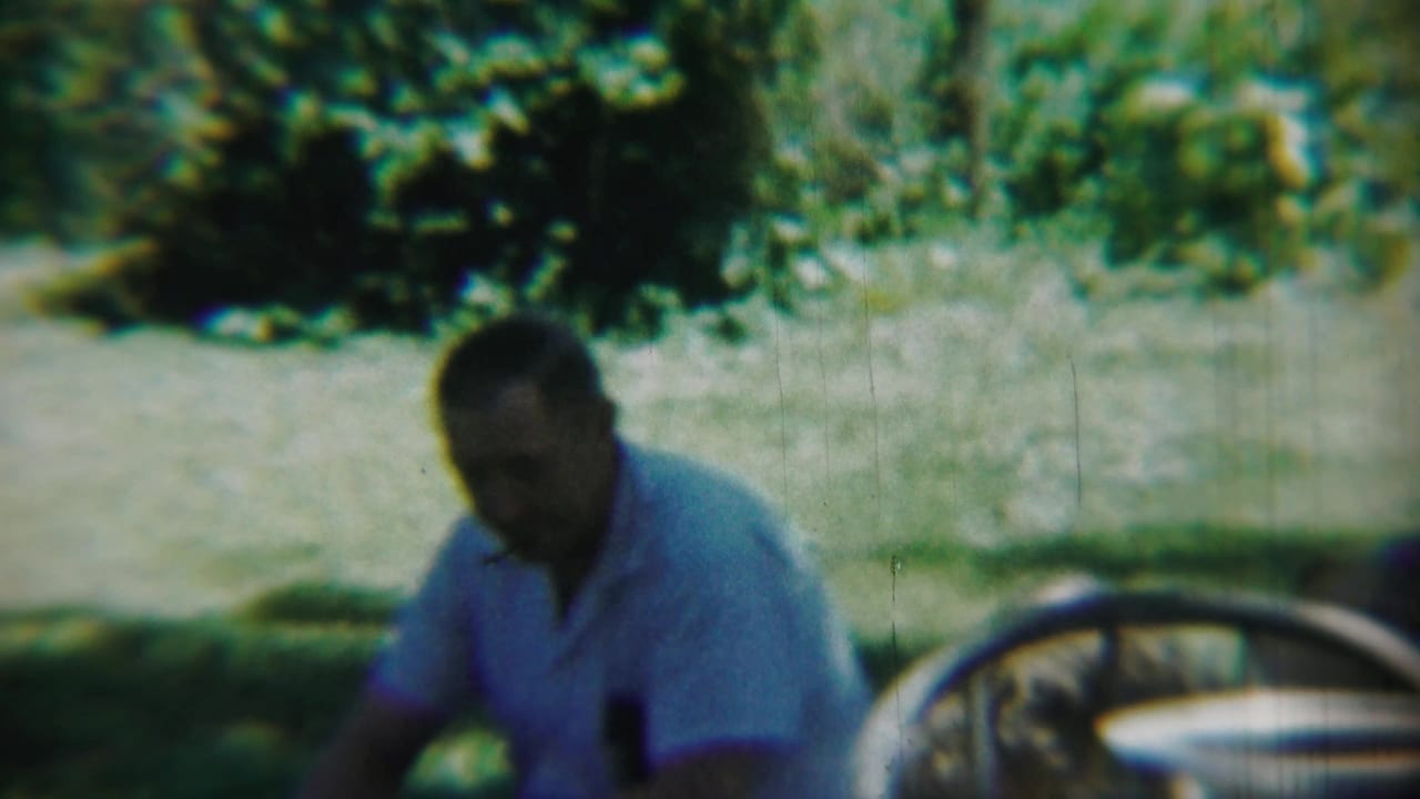 Chadron, Nebraska 1959 frame showing a man seated outdoors in a light blue shirt, with trees, grass, and a partial car wheel nearby.