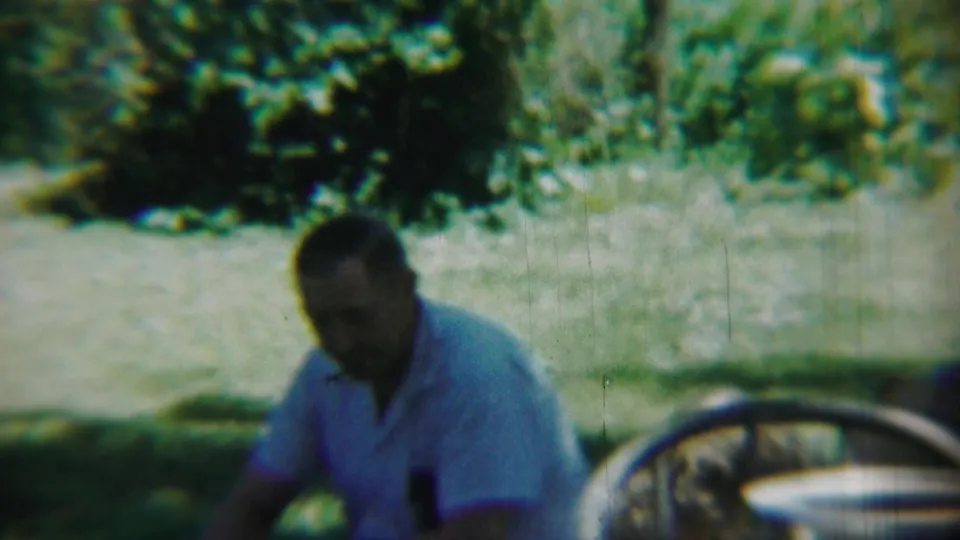 Chadron, Nebraska 1959 frame showing a man seated outdoors in a light blue shirt, with trees, grass, and a partial car wheel nearby.