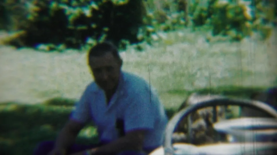 Soft 1959 8mm frame from Chadron, Nebraska with a seated man in a light shirt, trees behind him, and part of a vintage car wheel at right.