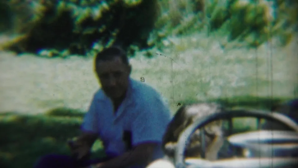Chadron, Nebraska 1959 home-movie frame showing the seated man outdoors near grass, trees, a pale path, and the edge of a car wheel.