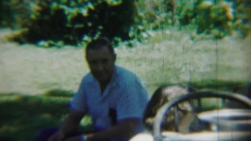 Adult man seated outdoors near a pale vintage car in Chadron, Nebraska home-movie footage from 1959.