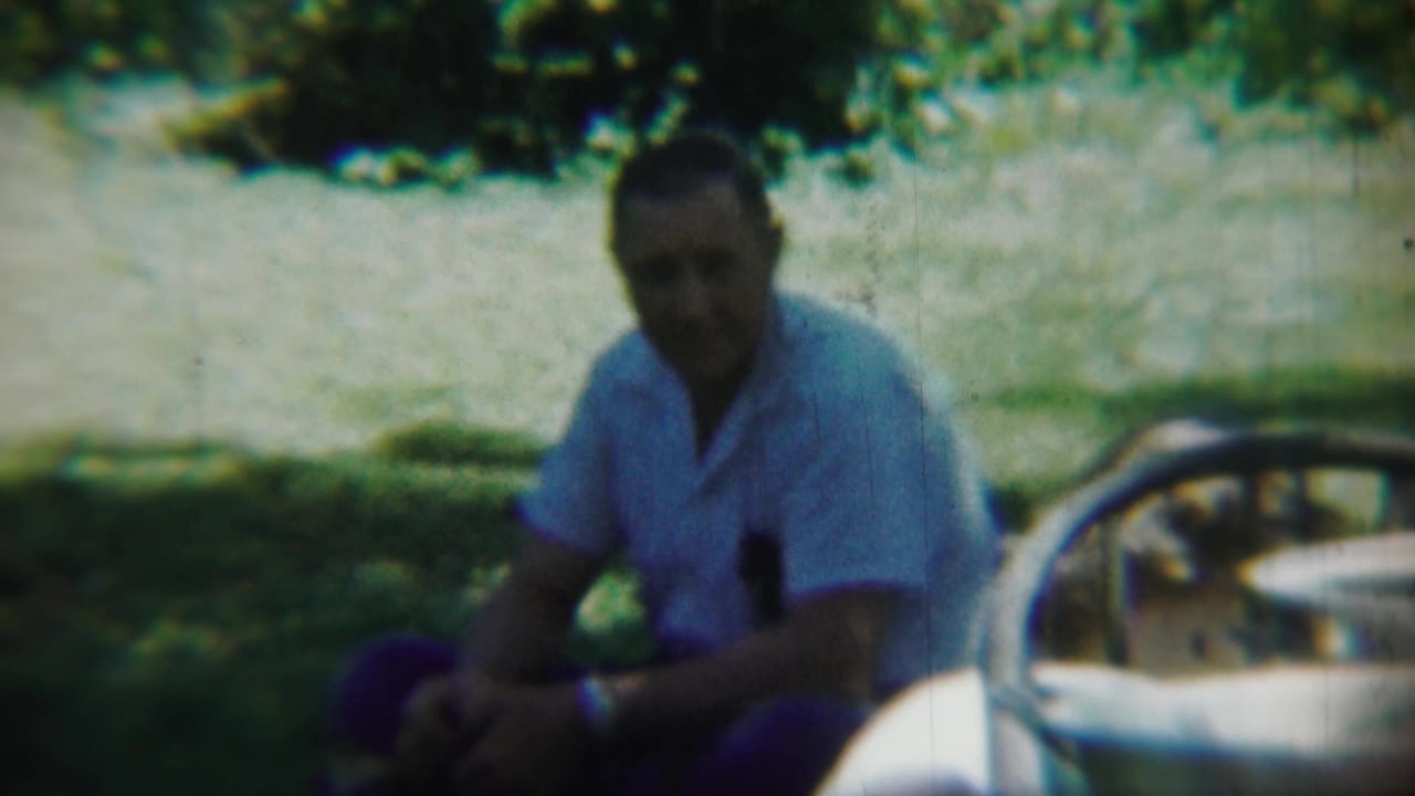 Man seated beside a light-colored car in a shaded grassy area.