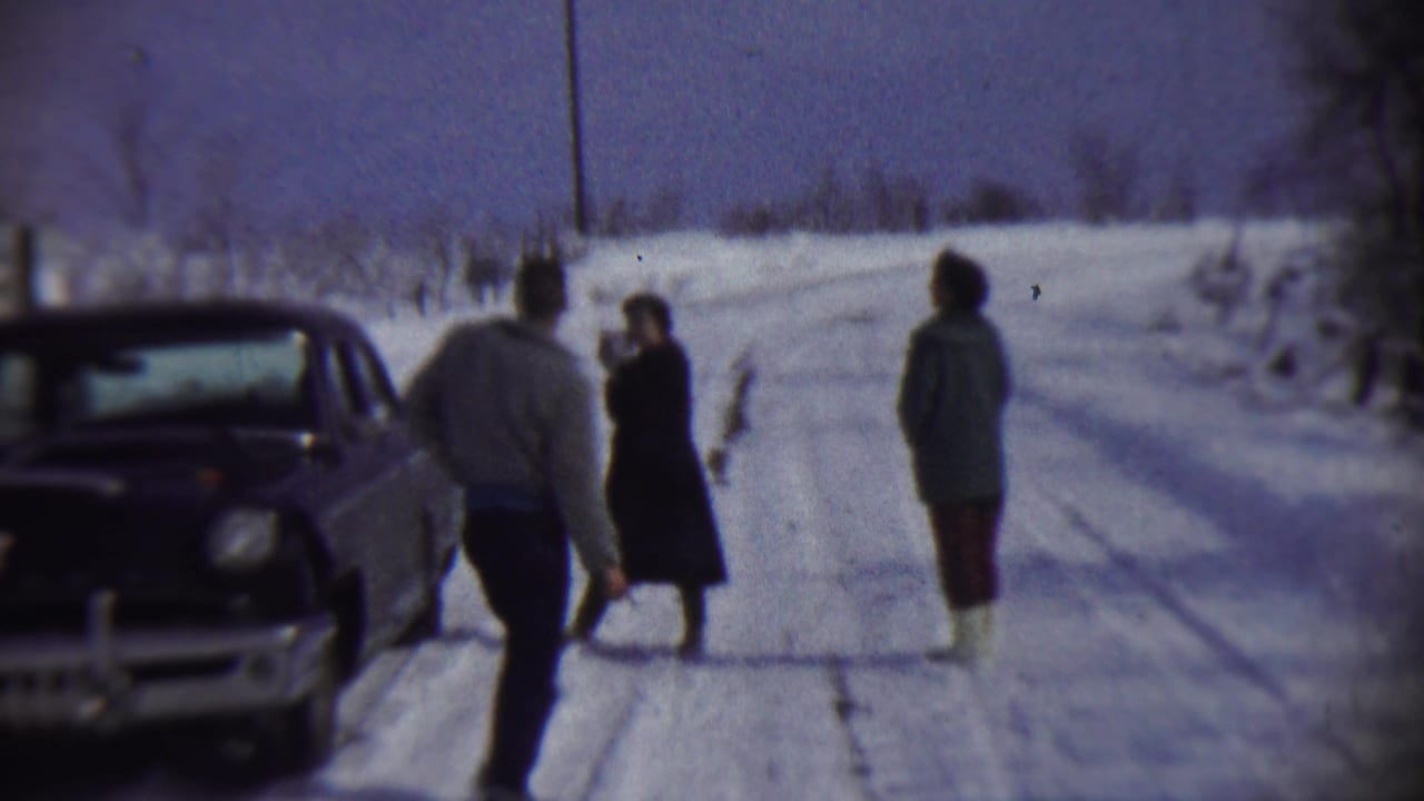 Three bundled people stand on a snowy rural road beside a parked vintage car.