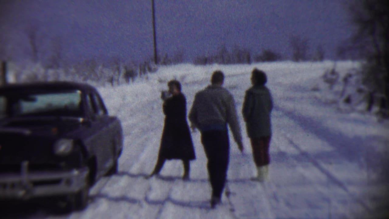 1959 Chadron, Nebraska frame with a woman holding a camera near a vintage car while two bundled companions move along the snowy road.