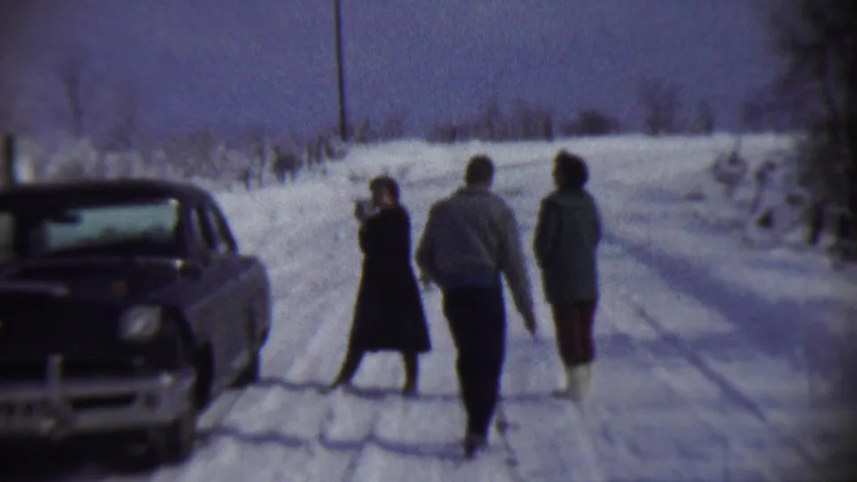 1959 Chadron, Nebraska frame with a woman holding a camera near a vintage car while two bundled companions move along the snowy road.