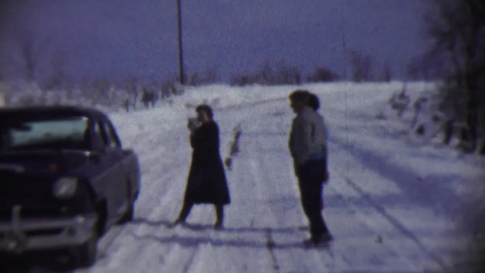 1959 Chadron, Nebraska home-movie frame showing two bundled people on a snow-covered road, with a parked vintage car partly visible at left.