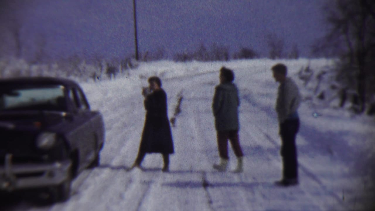 Three people stand on a snowy rural road beside a dark vintage sedan.