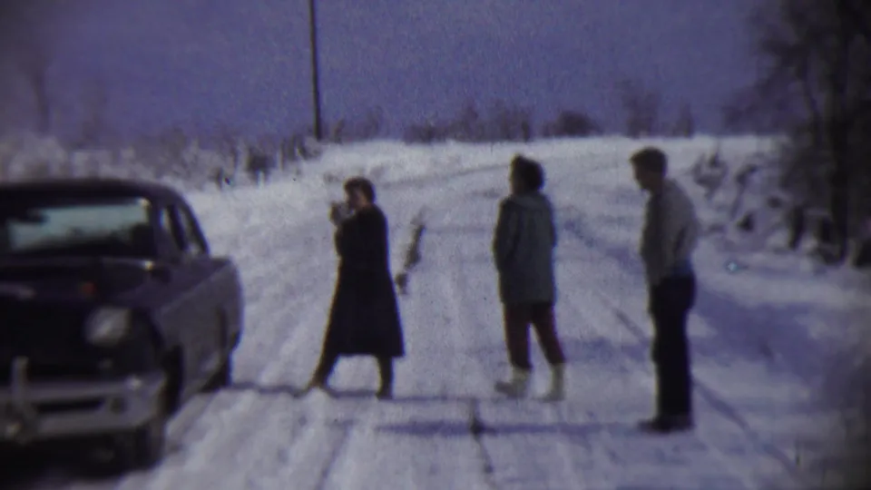 In 1959 Chadron, Nebraska, three adults stand on a snow-covered road near a dark 1950s sedan, with bare trees and brush behind them.