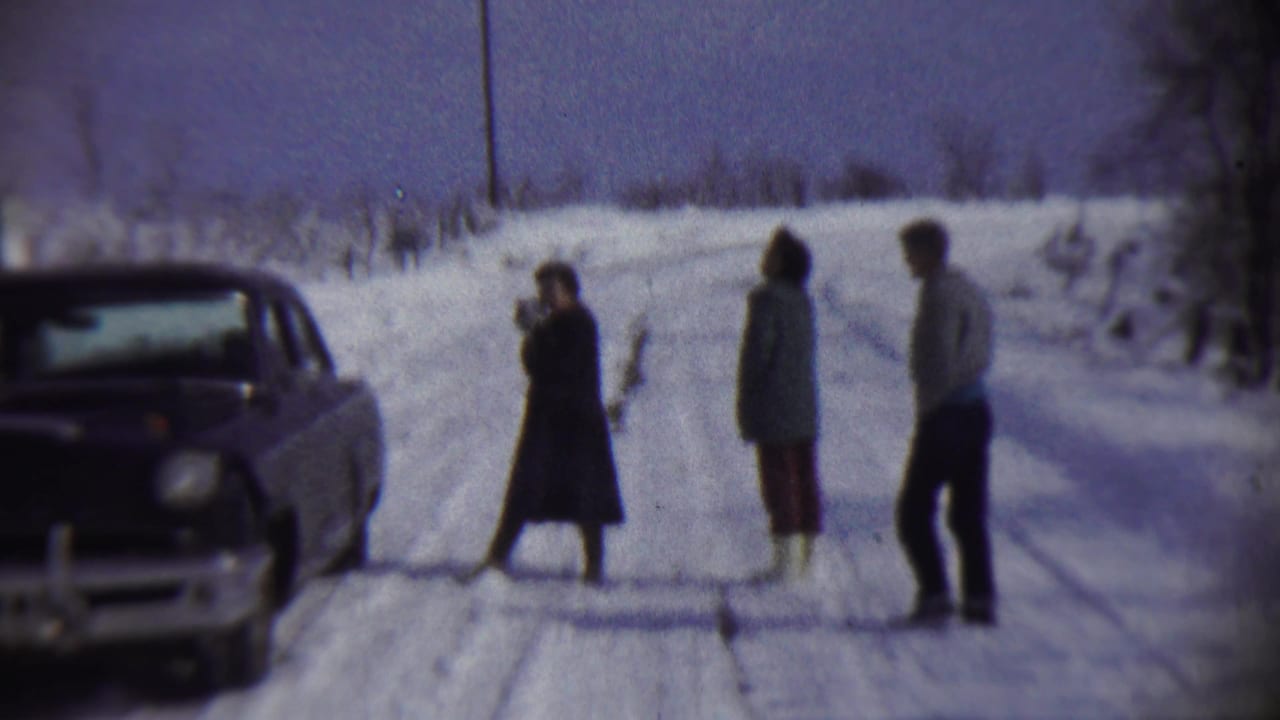 A woman in a dark coat appears to hold a camera while two others stand nearby on a snowy Chadron road beside a 1950s sedan.