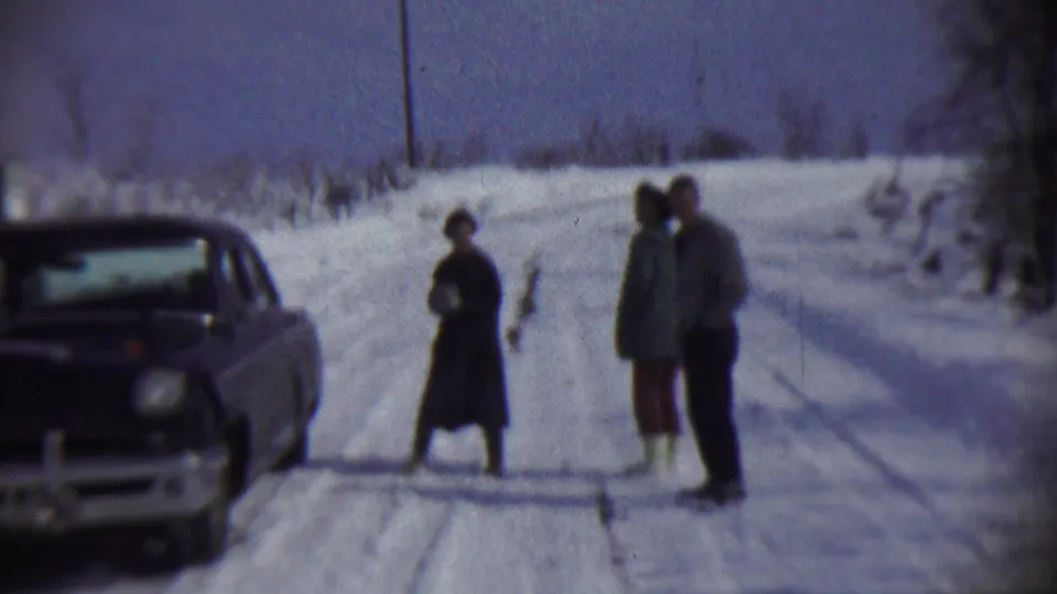 Three adults stand close together on a snow-covered rural road in Chadron, Nebraska, with the dark sedan partly visible at frame left.