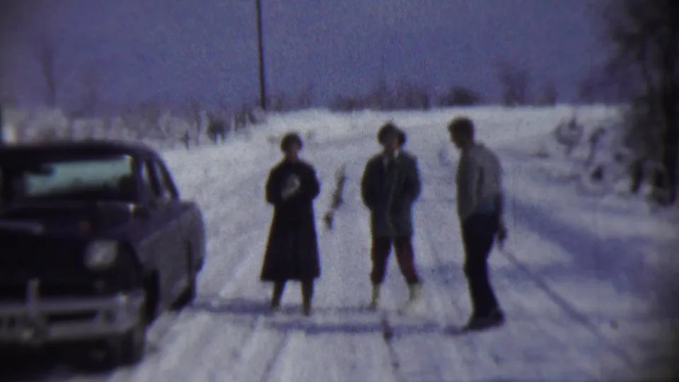 Three adults in winter clothing stand on a snowy rural road near a parked 1950s sedan, with open snowbanks and bare trees in the background.