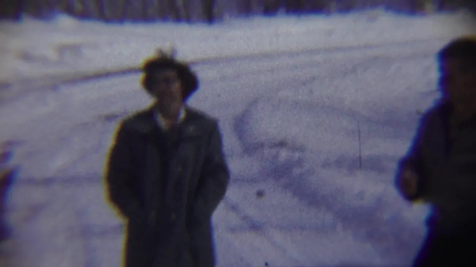 A bundled adult stands on the snowy roadside, with another person blurred in the foreground of the 1959 Chadron home-movie frame.