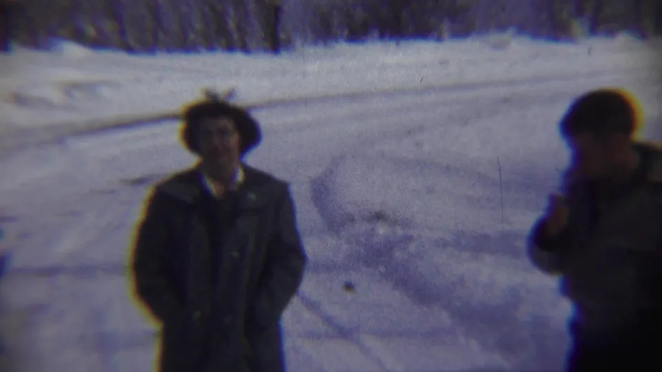 Two adults appear on a snowy rural road in Chadron, Nebraska, with one figure possibly lighting or holding a cigarette near his face.