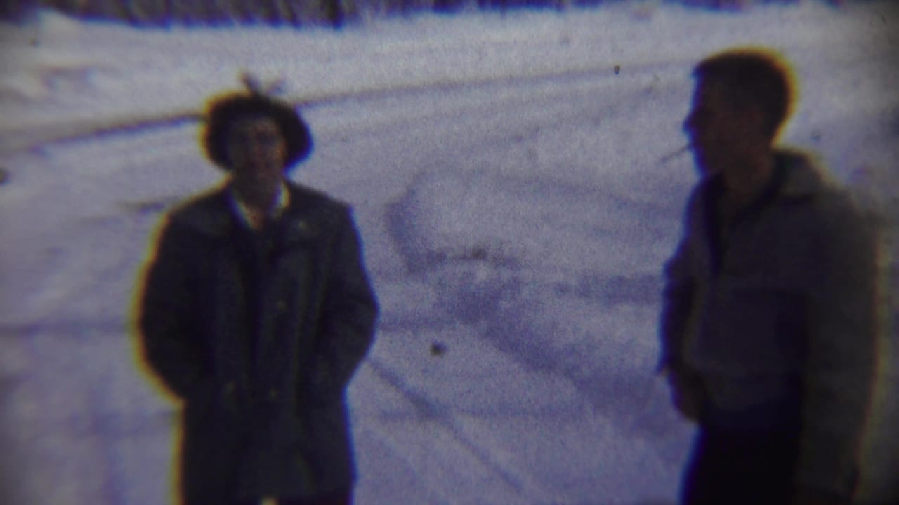 Two men stand in snow, with one in a light jacket smoking a cigarette.