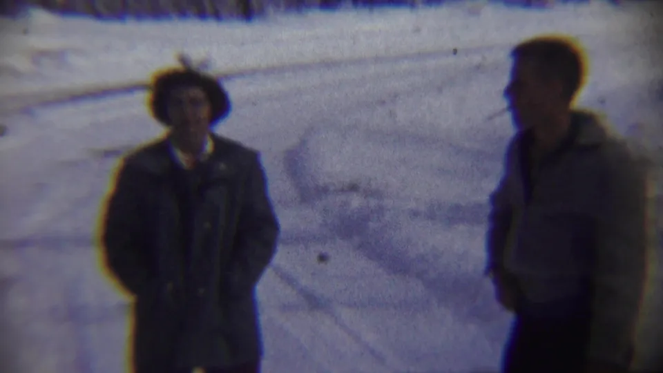 Two adult men stand in a snowy Nebraska landscape, one wearing a light jacket and smoking while the other faces the camera.
