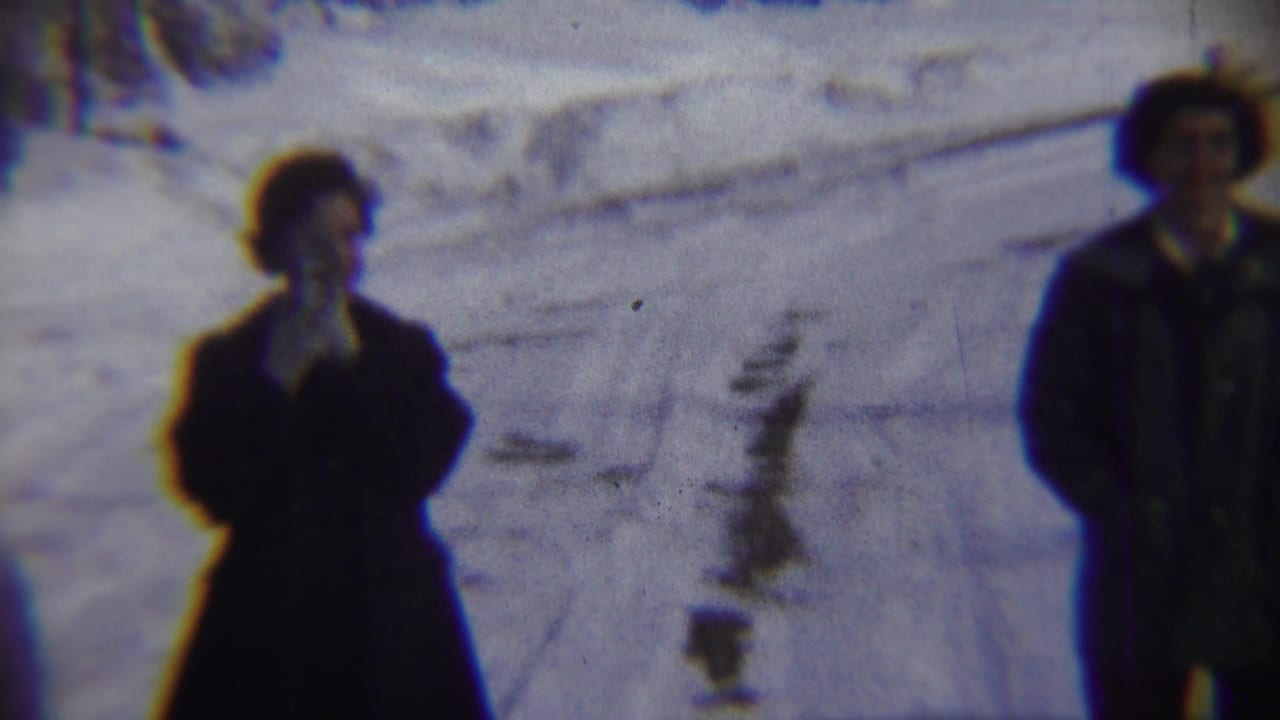 A woman in a dark coat appears to speak or hold a hand near her face while standing on a snowy Chadron street in 1959.