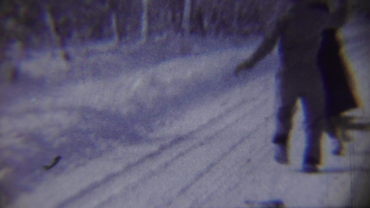 Two adults on a snowy road, one partly cropped, with trees and snowbanks behind them.