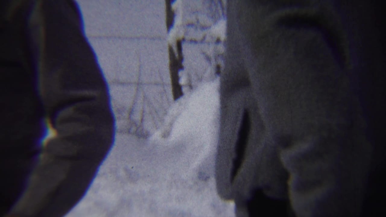 Two coat-wearing figures stand near a snow mound in an outdoor winter setting.