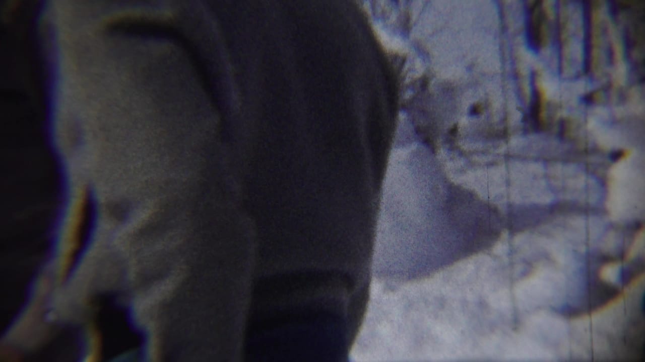 A bundled figure fills the foreground beside snow and a building in winter light.
