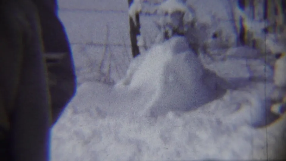 Two adults in winter coats stand near a deep snow mound in 1959 home-movie footage from Chadron, Nebraska.