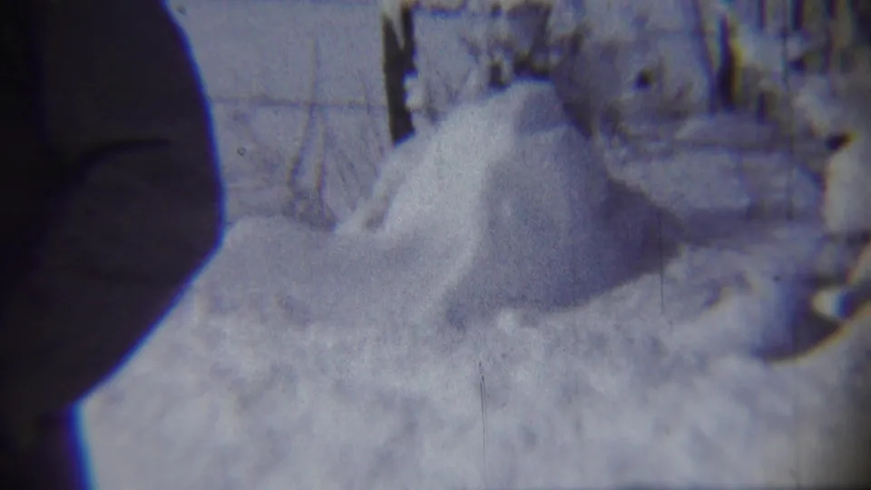 1959 8mm frame from Chadron, Nebraska, showing a deep snowdrift in a front yard with a dark edge silhouette and soft house or fence lines behind it.