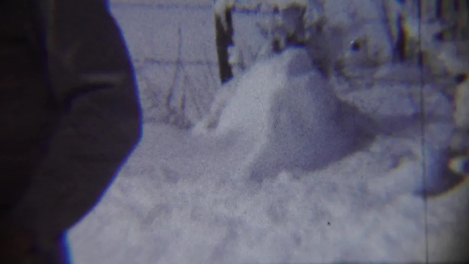 1959 Chadron, Nebraska home-movie frame showing a snowy yard and high drift, with a dark out-of-focus edge shape partly obscuring the view.