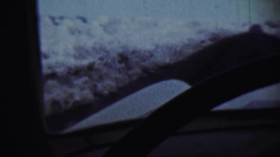 Home-movie frame from 1959 showing a winter landscape through a vehicle windshield near Chadron, Nebraska, with dashboard and wipers in shadow.