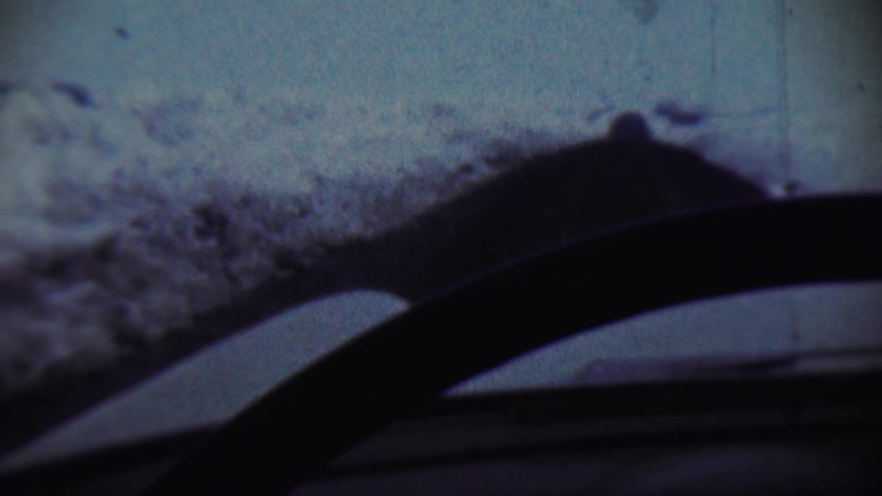 Snowy blue-toned view through a windshield with a dark curved car interior edge in the foreground.