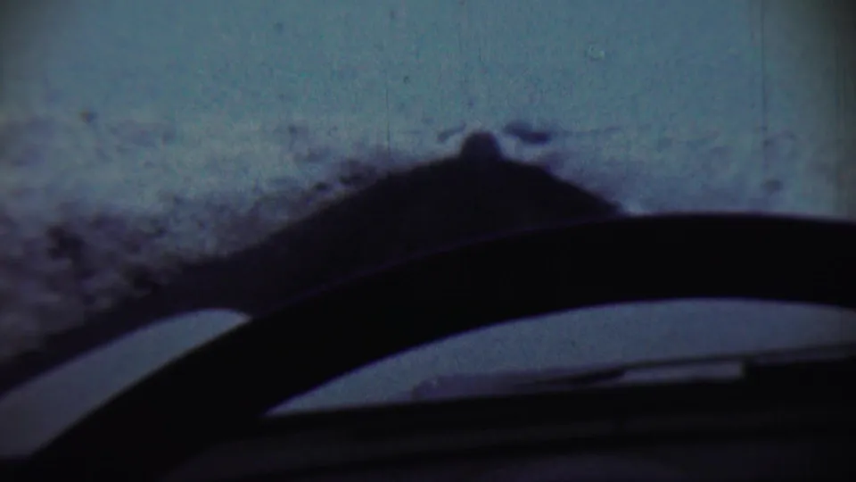 1959 home-movie still showing a low windshield view of winter snow, with the vehicle interior silhouetted across the foreground.