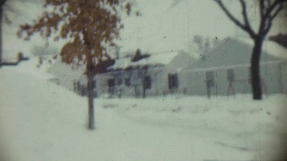 Soft 8mm frame from Chadron, Nebraska showing snow-covered yards, a small tree, bare trunks, and nearby houses.