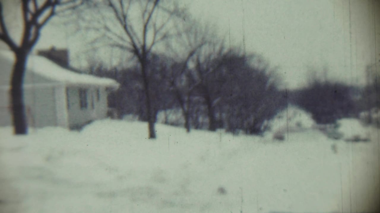 Snow-covered residential street with a house, bare trees, and deep drifts.