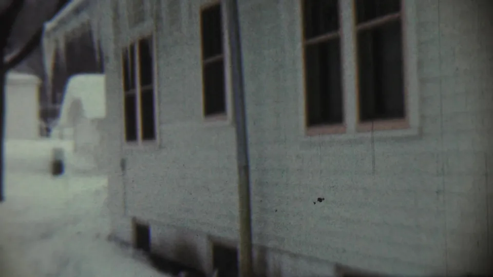 1959 Chadron, Nebraska frame with a close handheld view of white siding, dark window frames, and snow along the house.