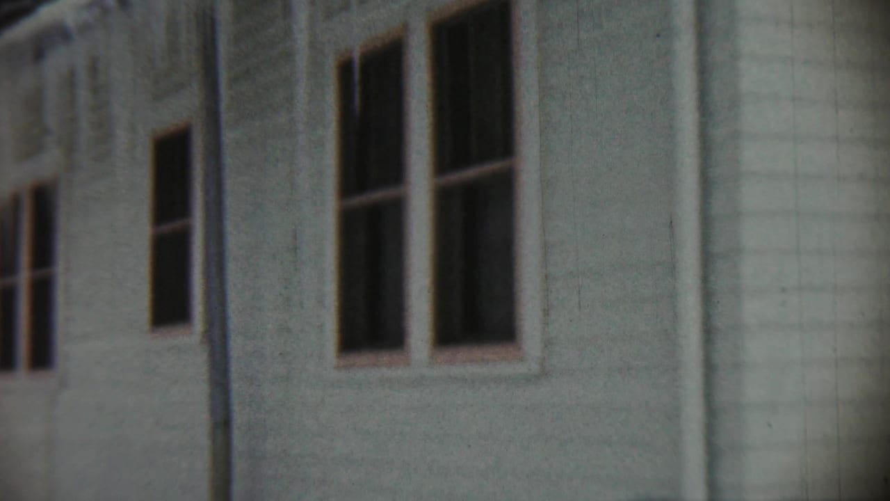 1959 Chadron home-movie frame showing a pale weathered house exterior with several dark window openings and vertical trim.