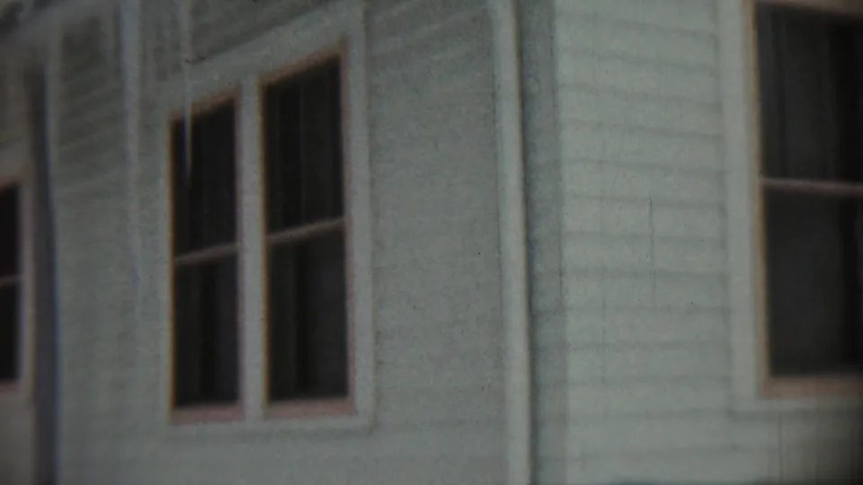 Close 8mm frame of a light-colored Chadron house exterior, with paired dark windows and worn siding texture visible.